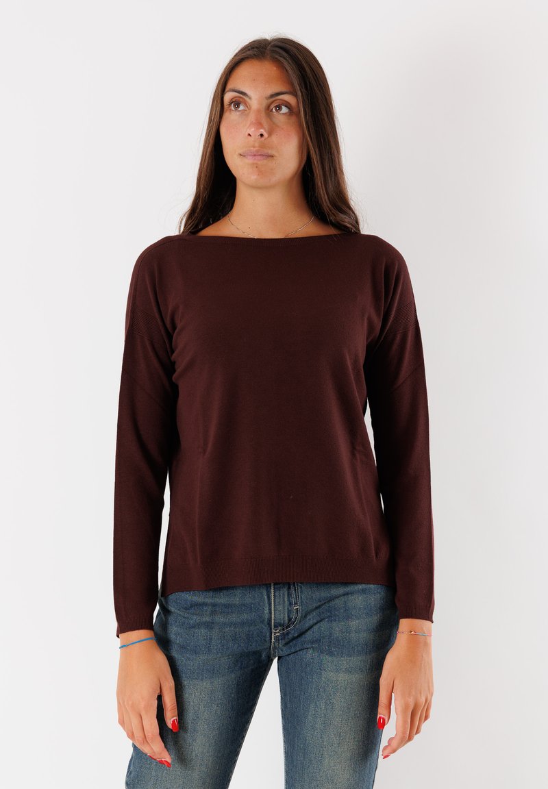 Maglione lungo marrone a maniche lunghe con scollatura ampia, realizzato in tessuto lavorato a maglia. Abbinato a un paio di jeans in denim blu, il modello si trova davanti a uno sfondo bianco.