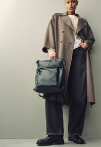 Schwarzer Leder-Rucksack mit Reißverschlussfächern, einseitig getragen, kombiniert mit einem übergroßen braunen Mantel, einem cremfarbenen Pullover und dunklen Jeans.