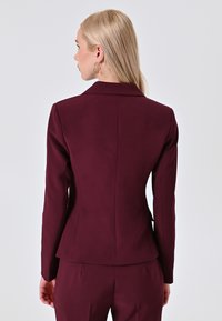 Blazer fitted color borgogna con maniche lunghe, colletto a revers e dettagli di cucitura in vista. Texture liscia, design sartoriale e silhouette strutturata.