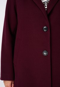 Gros plan sur une personne portant un manteau bordeaux avec deux boutons noirs sur une chemise rayée avec des détails en dentelle.