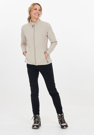 Kvinna som ler, stående med händerna i fickorna, iklädd beigerandig zip-up fleecejacka, svarta byxor och vandringsskor på vit bakgrund.