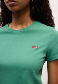 Femme portant un t-shirt vert uni Levi's avec un petit logo rouge Levi's sur la poitrine gauche, montrant une partie de son cou et de ses lèvres.