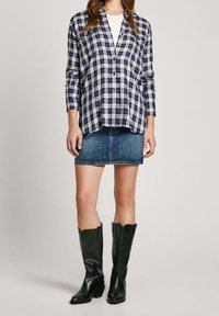 Kvinna iklädd en mörkblå rutig skjorta med knappar, en denimminikjol och svarta läderstövlar som når mitten av vaden mot en enfärgad bakgrund.