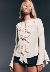 Blouse blanche à manches longues avec un devant à volants, des détails de boutons et des poignets évasés, associée à un short noir avec des accents métalliques.