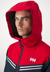 Rote und marineblaue Windbreaker-Jacke mit Kapuze, die weiße Streifen und ein Logo auf der Brust aufweist. Das Material wirkt glatt und leicht.