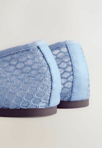 Chaussures à enfiler bleu clair avec tissu en mesh hexagonal ouvert et bordure solide, semelles plates marron, vues de dos sur une surface blanche.