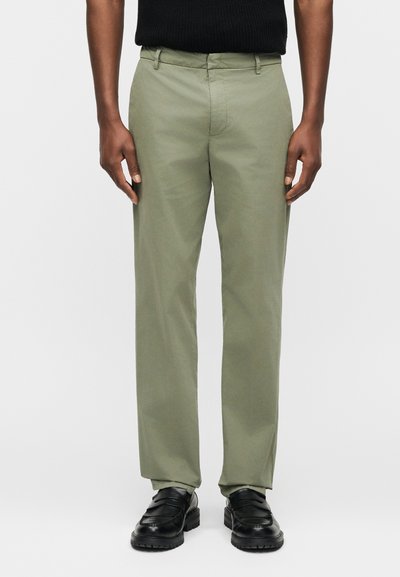 Άνδρας που φορά ελαιί πράσινο παντελόνι ευθείας γραμμής, μαύρα μοκασίνια και μαύρο αμάνικο τοπ, στέκεται μπροστά σε απλό φόντο.