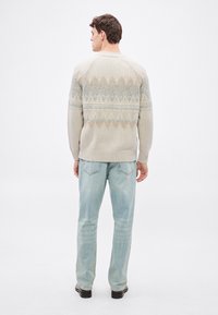 Beżowy sweter na drutach z geometrycznym wzorem w jasno niebieskim i brązowym kolorze. Połączony z jasno niebieskimi dżinsami i czarnymi butami. Widok od tyłu.