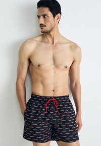 Shorts de baie negre cu un logo repetat "HUGO" în roșu și alb, cu un brâu elastic și detaliu cu siret roșu.