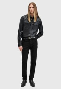 Camisa abotonada de cuero negro con dos bolsillos en el pecho, combinada con jeans negros y botas de tobillo negras, lucida por un modelo con cabello largo.