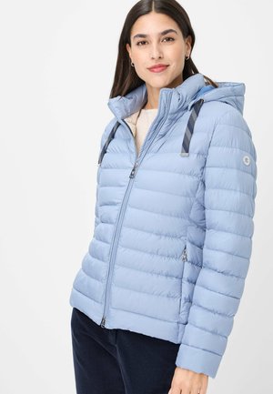 Frau trägt eine hellblaue, wattierte Steppjacke mit Kapuze und gestreiften Zugbändern, kombiniert mit dunkler Hose, stehend vor weißem Hintergrund.