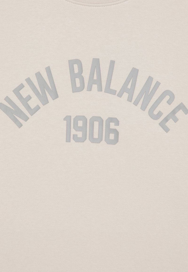 Sudadera beige con "NEW BALANCE" y "1906" en letras grises en relieve, cuello redondo y textura suave. Diseño minimalista.