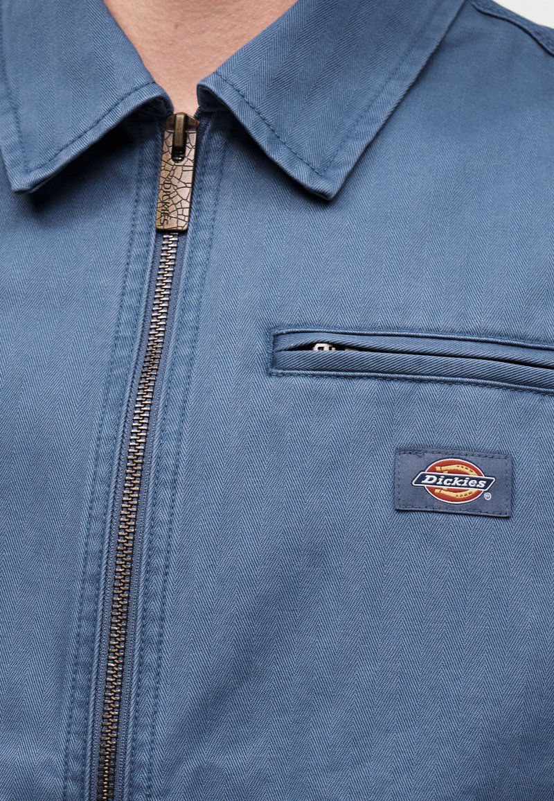 Close-up van een blauw werkjack met metalen ritssluiting, borstzak en Dickies merklogo patch aan de rechterkant.