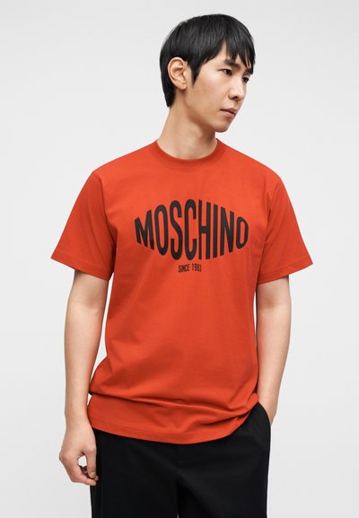 Κόκκινο βαμβακερό T-shirt με μεγάλο μαύρο λογότυπο "MOSCHINO" και κάτω από αυτό η επιγραφή "SINCE 1983". Με στρογγυλή λαιμόκοψη και κοντά μανίκια, απεικονίζεται σε ουδέτερο φόντο.