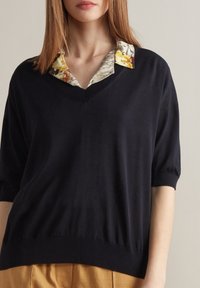 Tmavě námořnický pletený top s výstřihem do V, krátkými nabíranými rukávy a kontrastní květinovou košilí pod ním. Hladká textura, volný střih.