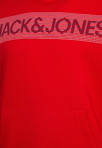 Sudadera roja con un gráfico de rayas horizontales en blanco y negro que incluye el texto "JACK & JONES". Cuenta con un bolsillo delantero y una textura de tela suave.