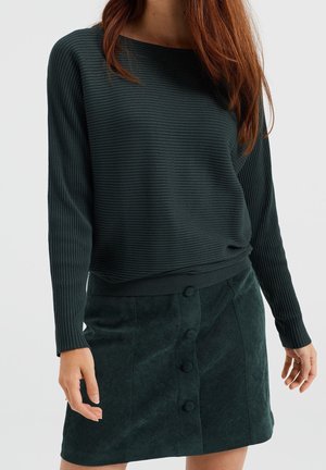 Vrouw met een donkergroen geribd shirt met lange mouwen en een bijpassende suède minirok met knopen aan de voorkant, staand met haar handen ontspannen langs haar zij.
