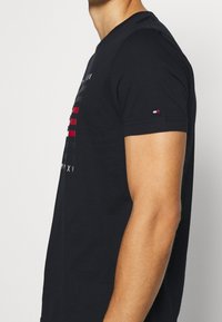 Camiseta negra de algodón con rayas horizontales en tonos rojos y grises, con un pequeño detalle de logo en la manga izquierda. Mangas cortas, cuello redondo.