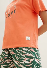 Korallefarbendes T-Shirt mit grauem "LOVE"-Text, kombiniert mit grüßen Hosen, die ein pink-weißes, abstraktes Blattmuster aufweisen. Etikett sichtbar am Saum des T-Shirts.