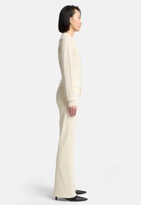 Cremefarvet langærmet skjorte kombineret med lysebeige viddebukser. Outfit'et fuldendes af sorte spidse sko. Ren, minimalistisk design.