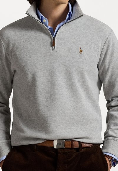 Άντρας φορεί ανοιχτό γκρι πουλόβερ με φερμουάρ μέχρι το τέταρτο πάνω από μπλε πουκάμισο, καφέ δερμάτινη ζώνη, σκούρα παντελόνια και ρολόι στον αριστερό καρπό.