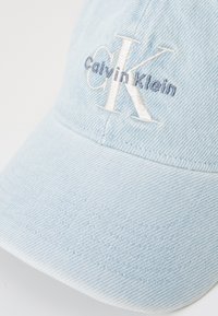 Gaiši zils džinsa cepures ar izšūtu "Calvin Klein" logotipu baltā un pelēkā krāsā. Iespējojama struktūras mala un teksturēta auduma virsma.