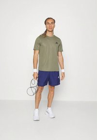 T-shirt atlético verde-oliva, calções azuis escuros, ténis brancos, segurando uma corda de pular. Design simples com o logótipo da Adidas na t-shirt.