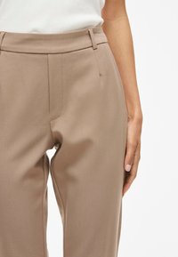 Beige skräddarsydda byxor tillverkade av ett mjukt tyg, med framfickor, sidofickor och midjehöjd som är medelhög. Enkel design utan synlig hårdvara.