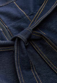 Dunkelblauer Denimstoff mit einem Knoten-Detail und kontrastierenden gelben Steppnähten entlang der Nähte, die die Textur und Handwerkskunst hervorheben.
