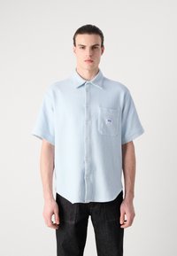 Camicia azzurra a maniche corte con bottone, in tessuto a costine, dotata di taschino sul petto con logo e orlo arrotondato. Abbinata a pantaloni scuri.