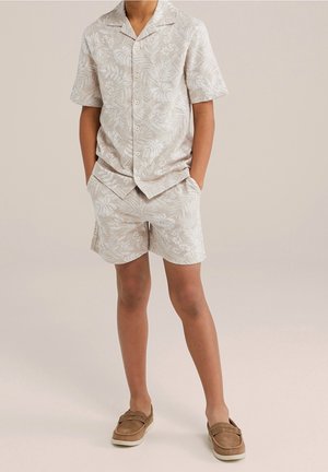 Jongen die een beige overhemd met korte mouwen en bijpassende short draagt, beide met een wit bladpatroon, tan instapschoenen en handen in de zakken.