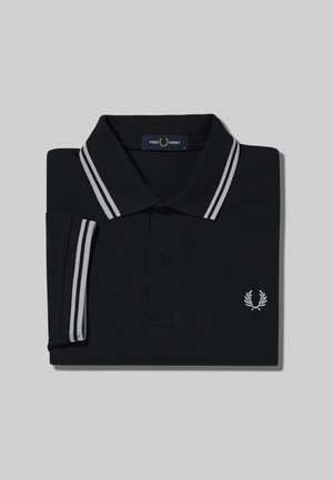 Polo negro de Fred Perry con dobles rayas blancas en el cuello y las mangas, doblado cuidadosamente sobre un fondo gris.