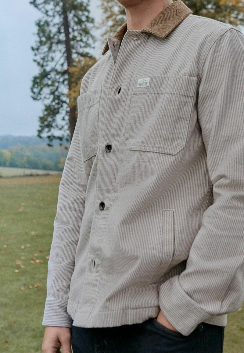 Homme portant une veste en velours côtelé beige avec un col marron, debout à l'extérieur, main dans la poche, arbres et champ herbeux en arrière-plan.
