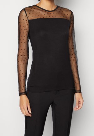 Femme portant un haut noir transparent à pois à manches longues avec un tissu noir uni sur la poitrine et le torse, associé à un pantalon noir.