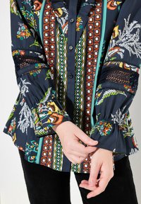 Blusa floreale blu navy con motivi vivaci, maniche a volant e chiusura con bottoni. Decorazioni includono inserti in pizzo sulle maniche.