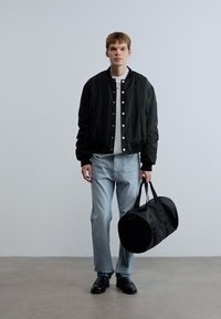 Μαύρο bomber μπουφάν με λευκά κουμπιά, γκρι t-shirt, ανοιχτό μπλε τζιν και μαύρα παπούτσια. Κρατάει μια στρογγυλή μαύρη τσάντα duffle.