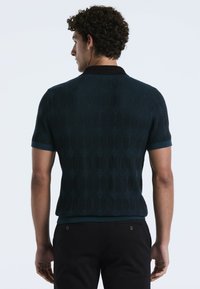 Man met krullend haar, gekleed in een marineblauw gemusterd poloshirt met korte mouwen en zwarte broek, met de rug naar de camera tegen een effen achtergrond.