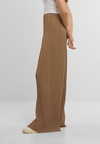 Bruine geplooide rok van zacht materiaal, met een hoge taille en vloeiende silhouet, gedragen met witte sneakers. Zijaanzicht weergegeven.