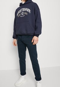 Tmavě modrá mikina s kapucí a přední kapsou, bílým potiskem, kombinovaná s tmavými kalhotami a bílými teniskami s texturovanou podrážkou a světlými detaily.