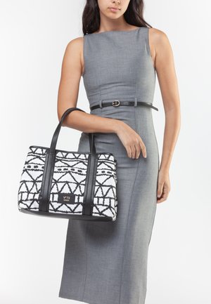 Donna con abito grigio senza maniche che tiene una borsa tote con motivo geometrico bianco e nero, manici neri e una piccola patch con logo.