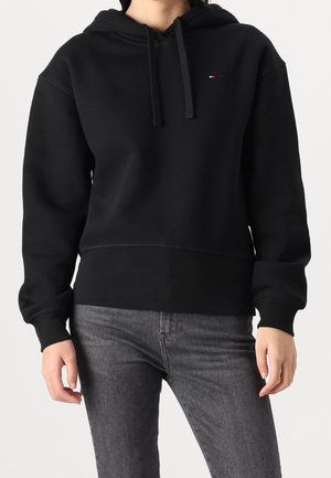 Personne portant un sweat à capuche noir avec cordons et un petit logo sur la poitrine, associé à un jean noir délavé, sur un fond uni.