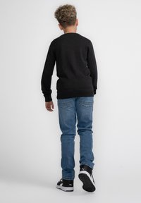 Schwarzer langärmeliger Pullover, blaue Denim-Jeans und schwarze Sneakers mit weißen Akzenten. Lässiger Schnitt mit sichtbaren Nähten und Design-Details auf der Rückseite.