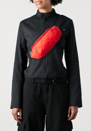Sac banane matelassé rouge porté en diagonale sur une veste noire, avec une texture lisse, fermeture éclair et sangle réglable.