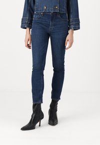 Tmavomodré denimové džínsy so slim fit strihom, vysokým pásom a jemnými švovými detailami, doplnené čiernymi čižmami s špicatou špičkou.