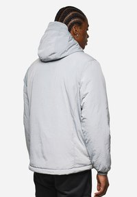 Giacca puffer grigia chiara con superficie texturizzata e cappuccio. Le caratteristiche includono polsini elastici e una vestibilità leggermente ampia.