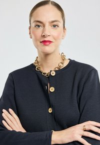 Gilet tricoté bleu marine avec un col rond, des boutons en bois et un détail en trou de serrure. Associé à un collier en chaîne dorée chunky.