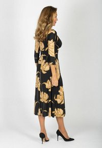 Mustakukkainen mekko, jossa on kolme neljännesosaa hihat, suurten vaalean beigejen kukkakuviolien kanssa ja leikkisä siluetti, jota täydentävät mustat korkokengät.