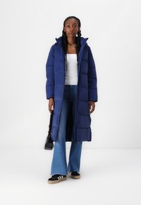 Palton lung pufos de culoare bleumarin cu un design quilted vertical, guler înalt și închidere cu fermoar. Asortat cu blugi evazați de culoare albastră deschisă și adidași negri.