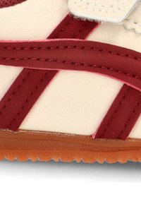 Zapatilla beige con acentos rojos, con un acabado suave, diseño en capas y una suela de goma texturizada. Cierre de velcro para facilitar su uso.