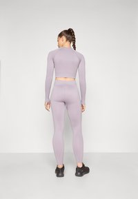 Svijetloljubičasti sportski crop top i tajice, izrađeni od glatkog materijala, prilagođenog dizajna, s dugim rukavima i visokim strukom. Crna obuća.